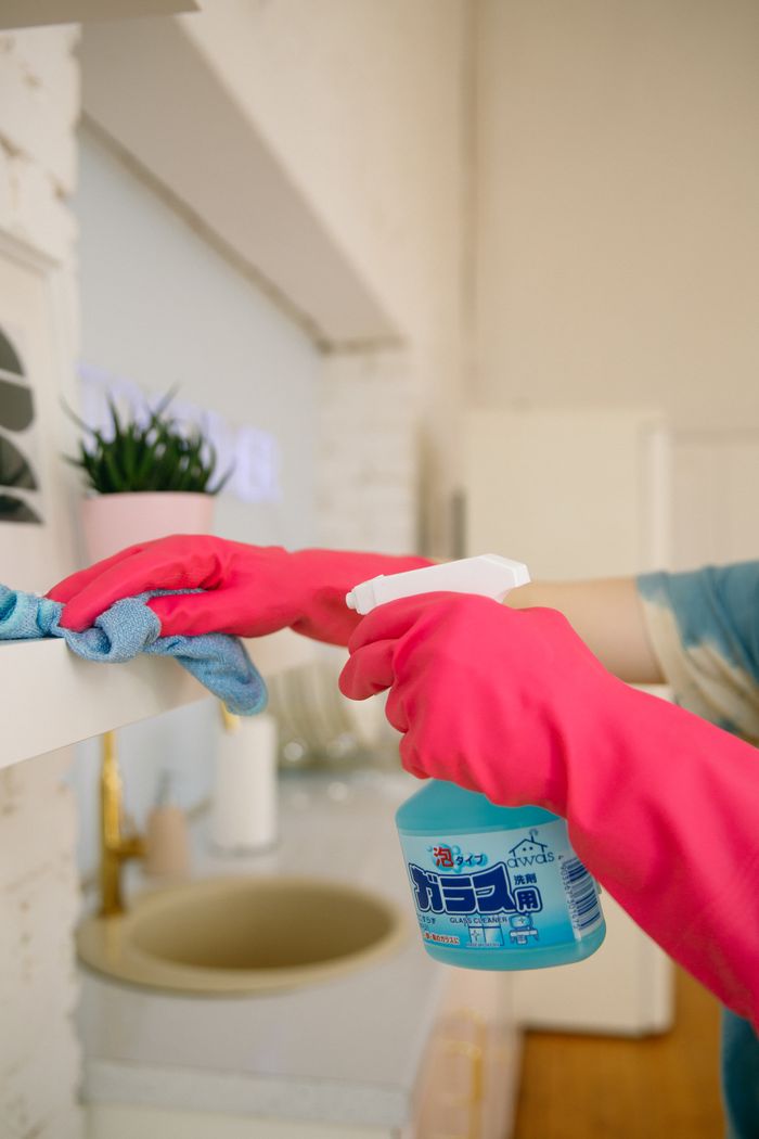 hero-02 Person using spray bottle and cloth for cleaning a kitchen shelf, ensuring hygiene and cleanliness.